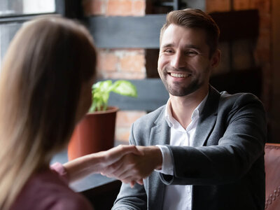 Job-Speed-Dating Ein Mann mit Bart und einem grauen Anzug sitzt lächelnd an einem Tisch und schüttelt einer Frau die Hand. Die Frau, deren Rücken zu sehen ist, hat lange blonde Haare und trägt ein burgunderfarbenes Oberteil. Beide scheinen sich in einem Büro oder Besprechungsraum zu befinden, der mit Pflanzen und modernen Möbeln ausgestattet ist.