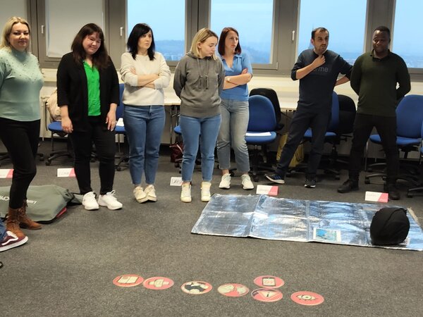 Kollegium fit für erste Hilfe Eine Gruppe von sieben Personen steht in einem Klassenzimmer und beobachtet eine Übungssituation zur Ersten Hilfe. Auf dem Boden liegt eine Rettungsdecke mit Symbolen für verschiedene Notfallmaßnahmen. Die Teilnehmer wirken konzentriert und aufmerksam.