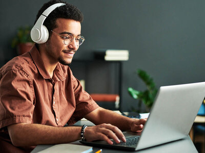 Ein Mann mit Brille und Kopfhörern sitzt an einem Schreibtisch und arbeitet an einem Laptop. Er trägt ein braunes Hemd und scheint konzentriert oder engagiert in seine Arbeit vertieft zu sein. Der Raum ist modern eingerichtet, mit einer Pflanze im Hintergrund und einem Regal mit Büchern und anderen Gegenständen. Der Raum ist gut beleuchtet und wirkt wie ein Büro oder ein Arbeitszimmer.
