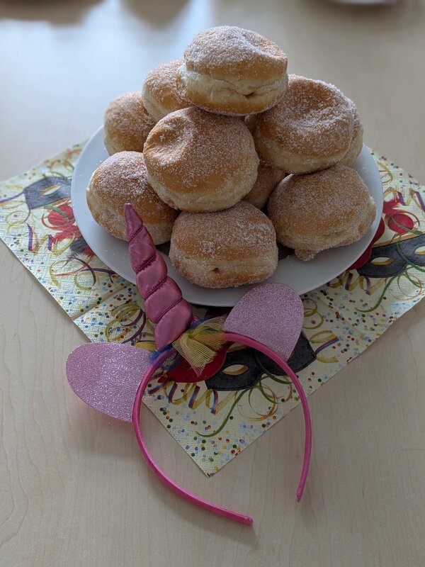 Faschingsdienstag an der Euro Akademie und den Euro-Schulen Dresden, Meißen und Riesa Ein Teller mit einer großen Portion Berliner (Krapfen) mit Puderzucker liegt auf einem farbenfrohen Karnevals-Servietten-Set. Daneben liegt ein pinker Haarreif mit einem Einhorn-Horn und glitzernden Ohren, passend zur Faschingsstimmung.
