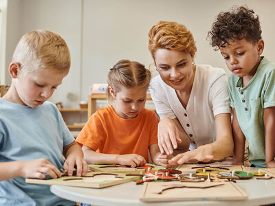 Teilnehmende eines Berufssprachkurses Frühpädagogik C1 arbeiten praxisnah mit Kindern in einer Lernsituation: Eine angehende Erzieherin unterstützt Kinder beim gemeinsamen Lösen eines Holzpuzzles in einem hellen Kita-Raum.