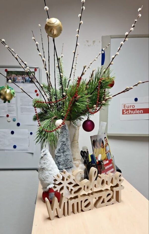 Gemeinsam auf Weihnachten freuen Ein weihnachtlich geschmückter Strauß aus Tannengrün und Weidenästen steht in einer weißen Vase auf einem Tisch. Davor steht eine Holzfigur, die aus den Worten "Schöne Winterzeit" und einer Schneeflocke besteht. Dahinter stehen zwei Wichtelfiguren aus Plüsch und eine Aufbewahrung mit Stiften, Scheren und Flyern von den Euro-Schulen.