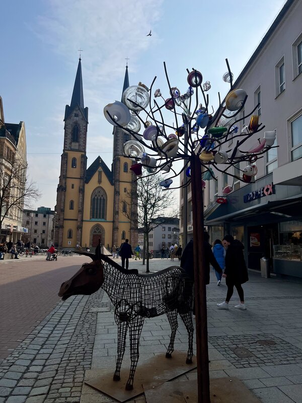 Abwechslung im Schulalltag: ein Kinobesuch Die Innenstadt von Hof. Zu sehen ist eine Gasse, an deren Ende die gelbe Marienkirche steht. Im Vordergrund stehen zwei Kunstwerke: Ein Esel aus Metall und daneben ein Baum aus Metall, an dessen Äste alte Lampenschirme aus Glas hängen.