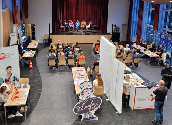 Lebhafte Podiumsdiskussion und Markt der Möglichkeiten Teilnehmende sitzen auf der Bühne und diskutieren, das Publikum hört zu, Informationsstände stehen bereit.