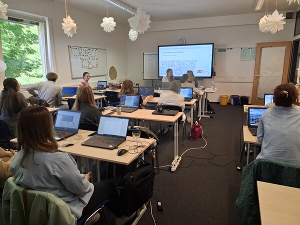 Teamtag der Euro-Schulen Berlin Brandenburg Mitarbeitende der Euro-Schulen sitzen zur Weiterbildung in einem großen Unterrichtsraum an Tischen, über ihnen hängen weiße Papiersterne, grauer Teppichboden ist ausgelegt. Vorn wird eine Präsentation am großen Bildschirm gezeigt.