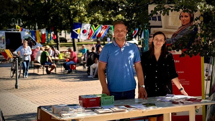 Interkulturelle Wochen im Landkreis Görlitz Ein Mann und eine Frau stehen an einem Infostand in einem Hof. Auf dem Tisch liegen Broschüren, dahinter steht ein Roll-up der Euro-Schulen. Im Hintergrund sieht man Menschen an Tischen sitzen, internationale Fahnen sind an einer Schnur aufgehängt. Es ist sonnig.