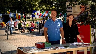 Interkulturelle Woche Ein Mann und eine Frau stehen an einem Infostand in einem Hof. Auf dem Tisch liegen Broschüren, dahinter steht ein Roll-up der Euro-Schulen. Im Hintergrund sieht man Menschen an Tischen sitzen, internationale Fahnen sind an einer Schnur aufgehängt. Es ist sonnig.