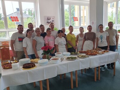 Teilnehmende stehen gemeinsam hinter der gedeckten langen Tafel, auf der ihre selbst zubereiteten Gerichte und Speisen stehen.