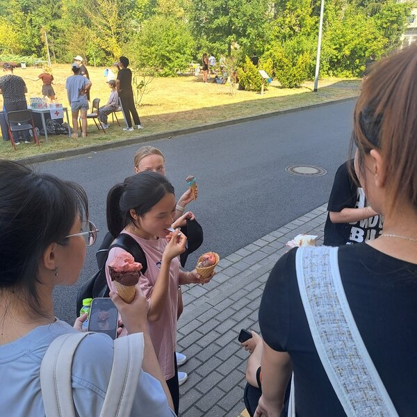 Sportfest – mal anders Mehrere Schülerinnen haben Waffeln mit verschiedenfarbigen Eiskugeln in ihrer Hand. Eine junge Frau hält in der anderen Hand ein Smartphone, auf dem das Foto einer Eiswaffel erkennbar ist. Im Hintergrund sieht man zwei Stationen des Sportfestes, wo weitere Personen stehen.