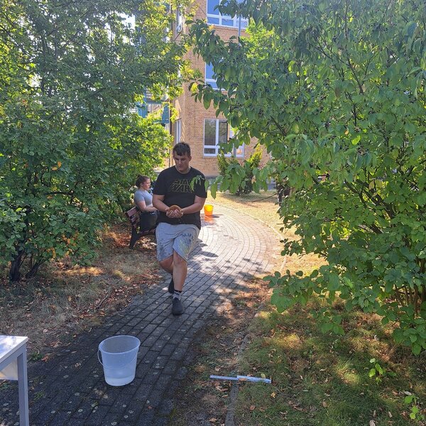 Sportfest – mal anders Ein Schüler läuft mit einem nassen Schwamm von einem mit Wasser gefüllten Eimer zu einem anderen mit Wasser gefüllten Eimer. Der Weg, auf dem er läuft, ist von Sträuchern umgeben. Im Hintergrund sitzt eine Schülerin auf einen Parkbank.