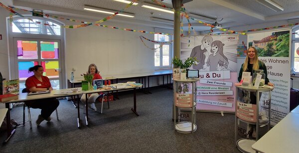 Hausmesse in Pößneck Ein heller Raum mit bunten Papiergirlanden und dekorierten Fenstern. Drei Frauen stehen bzw. sitzen an Info-Tischen, die zur Hausmesse dekoriert sind. Im Hintergrund: ein Roll-up mit der Aufschrift „Du & Du – Zwei Freunde“ der Hera Residenzen.