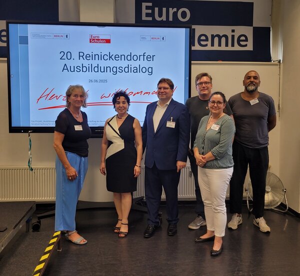 10 Jahre Reinickendorfer Ausbildungsdialog Mitglieder des Reinickendorfer Ausbildungsdialogs posieren für ein Gruppenfoto, im Hintergrund ein großer Bildschirm, auf dem die Teilnehmenden zum 20. RAD willkommen geheißen werden.