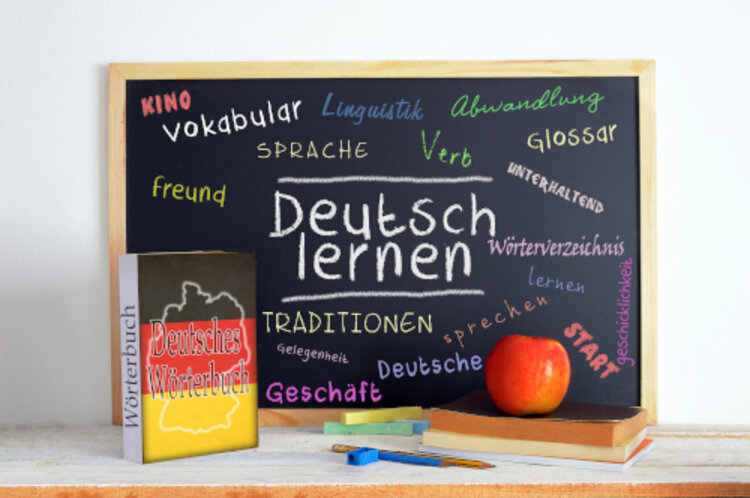 Deutsches Wörterbuch lehnt an einer Tafel, auf der "Deutsch lernen" und weitere deutsche Wörter stehen.