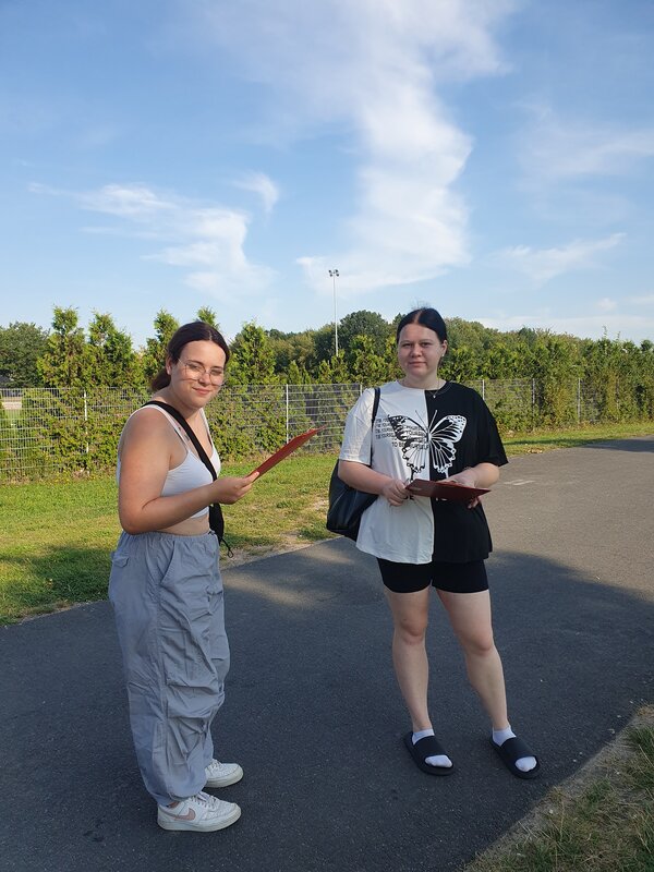 Naturvielfalt an der Goitzsche Zwei Schülerinnen in sommerlicher Kleidung stehen im Schatten auf einem asphaltierten Fahrradweg, dahinter befindet sich ein Sportplatz. Beide Mädchen halten eine Mappe in der Hand und haben eine Tasche umhängen.