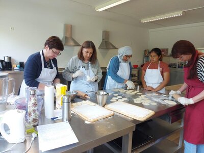 Eine Gruppe von Personen steht an einem Tisch und bereitet Teigtaschen zu. Sie rollen Teig aus und stechen Kreise aus, während andere die Füllung vorbereiten. Verschiedene Küchenutensilien stehen auf dem Tisch.