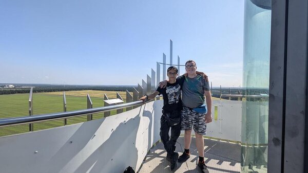 Abschlussfahrt: Erlebnispark Teichland Auf einem Aussichtsturm stehen zwei Jungs, die für das Foto die Arme umeinander gelegt haben. Im Hintergrund sind Windräder erkennbar, der Himmel ist wolkenlos.