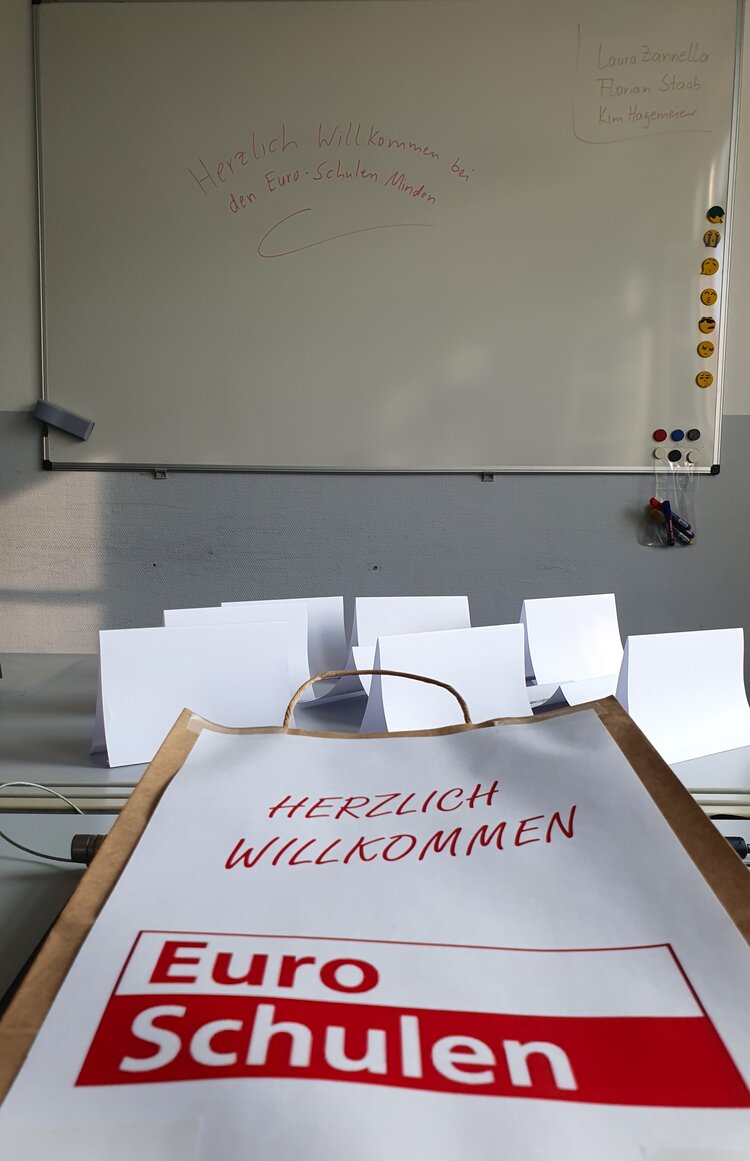 Zu sehen ein Prospekt mit dem roten Euro-Schulen Logo darauf, der auf einem Tisch liegt. I, Hintergrund ist eine weiße Tafel.