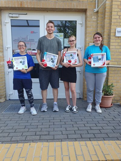 Vor der Tür der Euro-Schulen stehen drei zufrieden grinsende Schülerinnen und ein Schüler. Sieh halten individuell gestaltete, weiße Bilderrahmen in der Hand, sowie verschiedene kleine Geschenke.
