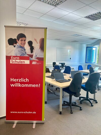 Unterrichtsraum Blick in einen Unterrichtsraum, in dem gepolsterte Schreibtischstühle und Tische stehen und ein großes Euro-Schulen-Roll up an der Seite steht.