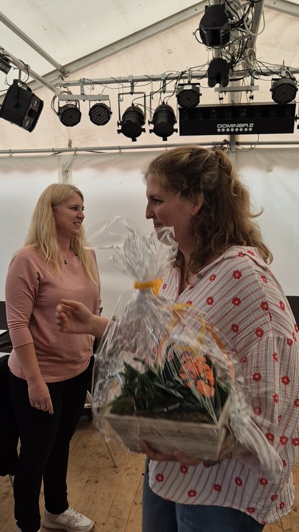 Grill & Chill bei den Euro-Schulen Niederrhein Eine scheidende Mitarbeiterin erhält zum Abschied eine Zimmerpflanze als Geschenk. Im Hintergrund steht (etwas unglücklich für die Szenerie) eine andere Mitarbeiterin, die nicht an diesem Gespräch beteiligt zu sein scheint. Das alles spielt sich in einem Festzelt ab, in dem auch eine professionelle Beleuchtungsanlage installiert ist.