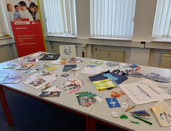 Netzwerken für die Zukunft auf der Weiterbildungsmesse Neuss Auf zwei großen Tischen liegen verschiedene Flyer und Werbegadgets unterschiedlicher Unternehmen. In der Ecke des Raumes, hinter den Tischen steht ein rotes Roll-up der Euro-Schulen Niederrhein.
