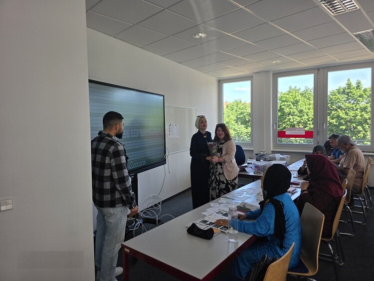 Vom Sprachkurs ins Berufsleben Die Teilnehmenden des Integrationskurses an den Euro-Schulen Dresden sitzen an Tischen, einige stehen vorn am Whiteboard mit der ehemaligen Teilnehmerin und überreichen ihr einen Blumenstrauß.