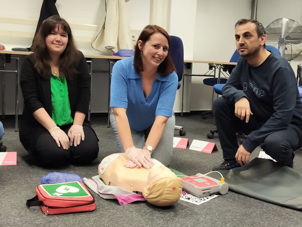 Kollegium fit für erste Hilfe Drei Personen nehmen an einem Erste-Hilfe-Kurs teil. Eine Frau in einem blauen Pullover demonstriert die Herz-Lungen-Wiederbelebung (HLW) an einer Übungspuppe, während zwei weitere Teilnehmer – eine Frau in einer schwarzen Jacke und ein Mann in einem dunklen Pullover – daneben knien und zuschauen. Vor ihnen liegt ein Erste-Hilfe-Set mit einem Defibrillator.