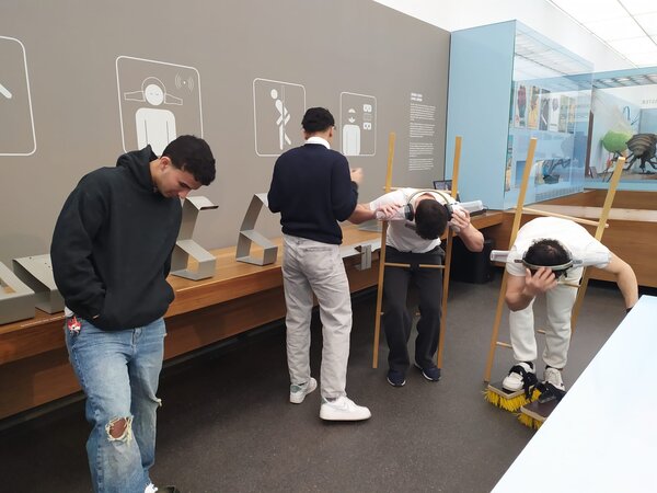 Sonniger Exkursionstag der Euro-Schulen Dresden: Spannende Einblicke in Wissenschaft und Kultur Mehrere Personen befinden sich in einem interaktiven Museum und probieren verschiedene Experimente aus. Zwei Personen haben sich in eine Vorrichtung gelehnt, die ihren Körper nach vorne kippen lässt, während eine andere Person Schuhe mit Bürsten trägt.