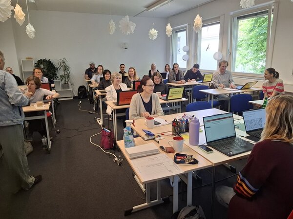 Teamtag der Euro-Schulen Berlin Brandenburg Mitarbeitende der Euro-Schulen sitzen zur Weiterbildung in einem großen Unterrichtsraum an Tischen, über ihnen hängen weiße Papiersterne, grauer Teppichboden ist ausgelegt.