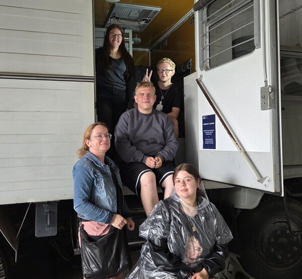 Exkursion zur Gedenkstätte Bautzen Fünf junge Menschen posieren vor einem weißen LKW mit offener Tür in einer Halle. Zwei Personen sitzen auf der Treppe zum Fahrzeug, eine Person sitzt vorne auf einem Hocker und trägt einen durchsichtigen Regencape. Zwei weitere Personen stehen seitlich und lächeln in die Kamera.