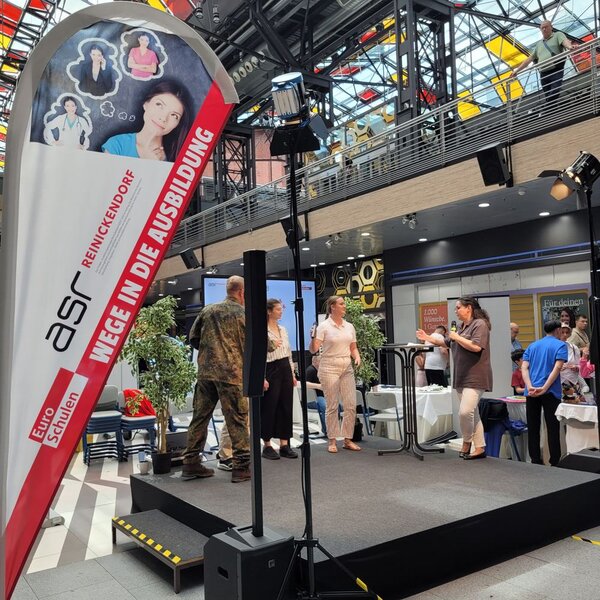 „Wege in die Ausbildung“ - Ein Tag voller Perspektiven für junge Menschen Die Vertreter*innen der beteiligten Ausbildungsunternehmen stellen auf der Bühne ihre Berufe vor. Links im Bild ist die Beach Flag der Euro-Schulen mit dem Schriftzug "ASR Reinickendorf - Wege in die Ausbildung" zu erkennen.