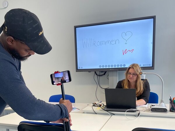 Mit KI zu barrierefreier Bildung – Teil 1 Mitarbeiterin sitzt vor dem Laptop am Schreibtisch und erklärt etwas, während der Kameramann sie aufnimmt.