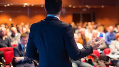 Ein Mann mit schwarzem Anzug steht vor Menschen in einem Saal und hält eine Rede.