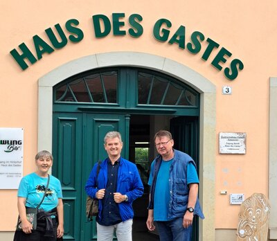 Drei Personen stehen vor dem Eingang des Gebäudes „Haus des Gastes“ und lächeln in die Kamera. Die Tür ist grün, darüber ist der Gebäudename in großen grünen Buchstaben angebracht.