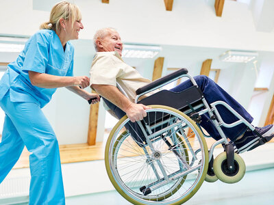 Eine Pflegekraft in blauer Uniform hilft einem älteren Mann in einem Rollstuhl. Die Pflegekraft steht hinter dem Rollstuhl und schiebt ihn, während der ältere Mann sich umdreht und lacht. Beide scheinen sich in einer Pflegeeinrichtung oder einem Krankenhaus zu befinden. Der Raum ist hell und freundlich gestaltet, mit großen Fenstern, die Tageslicht hereinlassen.