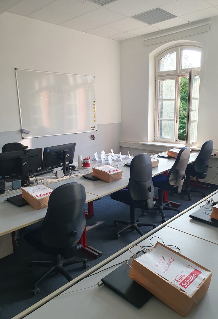 Zu sehen in Reihe gestellte Tische mit blauen Stühlen, auf denen Werbematerial der Euro-Schulen und Schreibwaren liegen.