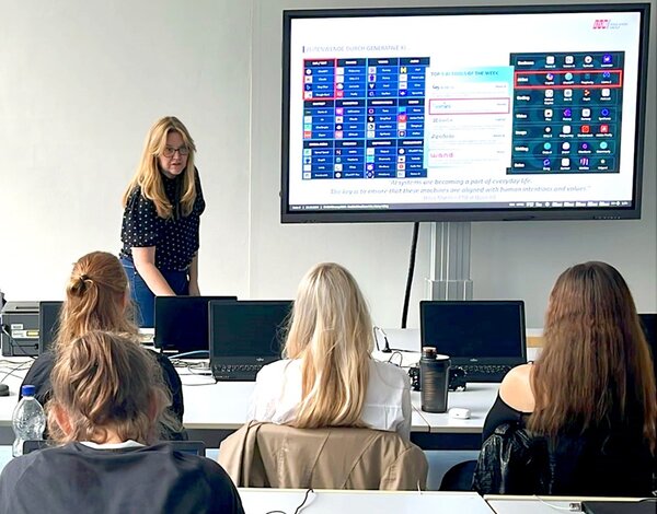 Workshopleiterin steht vorn im Klassenraum und referiert vor Schülerinnen und Schülern über Künstliche Intelligenz. 