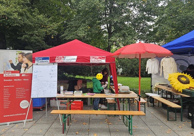Überdeckter Euro-Schulen Stand, mit allerlei Kuchen und Lehrmaterialien, einer Sitzbank und Tisch davor und einer Mitarbeiterin mit ihrem Kind.