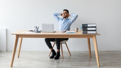 Zufrieden am Schreibtisch Ein lächelnder Mann sitzt entspannt auf einem Bürostuhl am Schreibtisch, vor einem Laptop, lehnt sich zurück und ist zufrieden mit seinem Job.