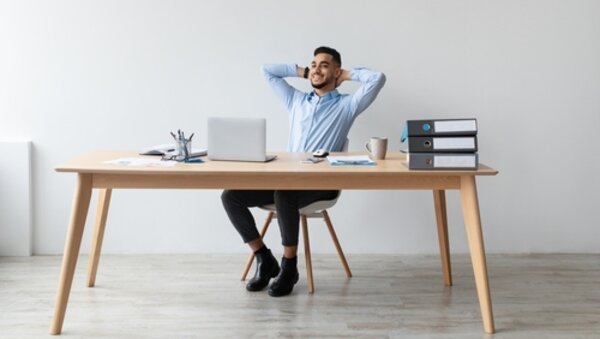 Hilfe bei der Berufswahl Ein lächelnder Mann sitzt entspannt auf einem Bürostuhl am Schreibtisch, vor einem Laptop, lehnt sich zurück und ist zufrieden mit seinem Job.