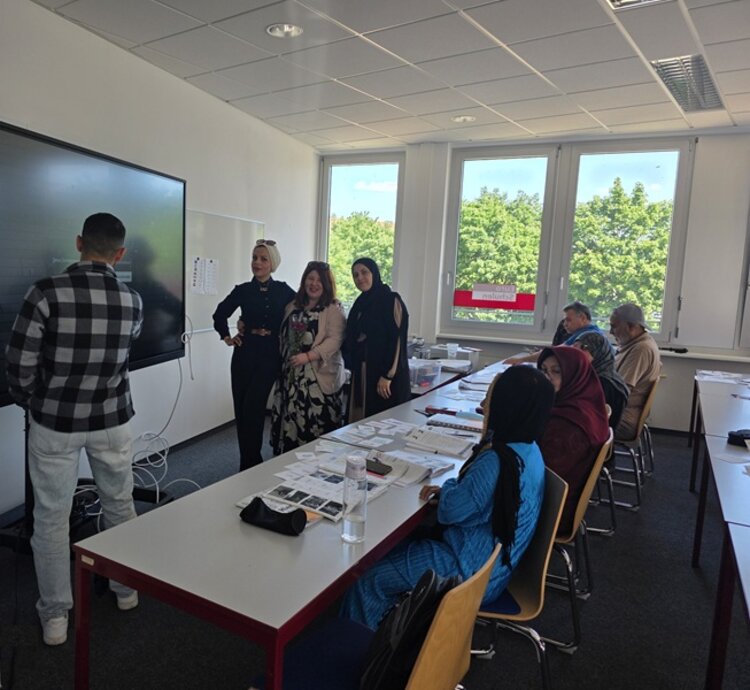 Vom Sprachkurs ins Berufsleben Die Teilnehmenden des Integrationskurses an den Euro-Schulen Dresden sitzen an Tischen, einige stehen vorn am Whiteboard mit der ehemaligen Teilnehmerin, die als Gast den Kurs besuchte, im Unterrichtsraum und posieren für ein Foto.