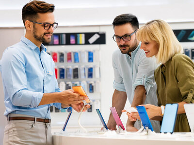 Drei Personen in einem Elektronikgeschäft, die sich über Smartphones unterhalten. Ein Verkäufer mit Brille und blauem Hemd zeigt einem Paar ein orangefarbenes Smartphone. Im Hintergrund sind verschiedene Smartphone-Modelle in ihren Verpackungen an der Wand zu sehen. Das Paar scheint sich für die Auswahl der Smartphones zu interessieren.
