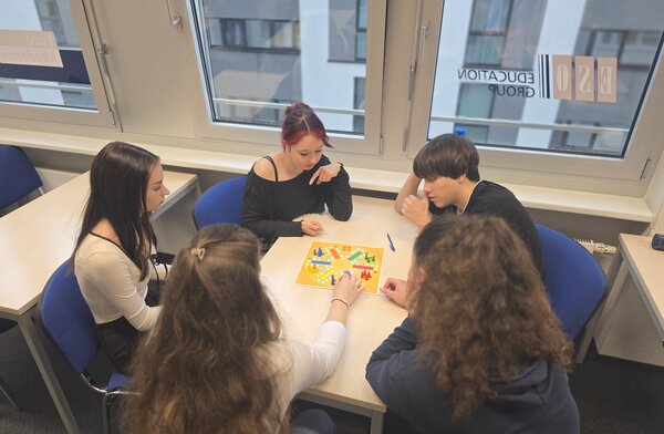 Spielen, Sprechen, Spaß haben – Kreative Gesprächsanlässe im Jugendintegrationskurs Fünf junge Erwachsene sitzen gemeinsam an einem Tisch und spielen „Mensch ärgere dich nicht“. Alle sind in das Gesellschaftsspiel vertieft.