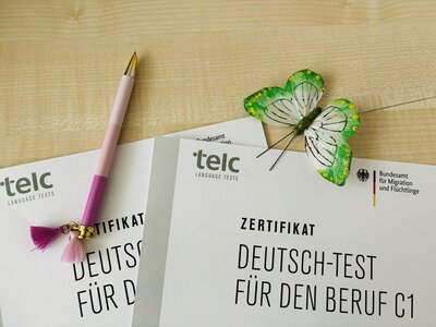 Zwei Zertifikate zum Deutsch-Test mit Zielsprachniveau C1, einem Kugelschreiber und einem Deko-Schmetterling sind abgebildet.