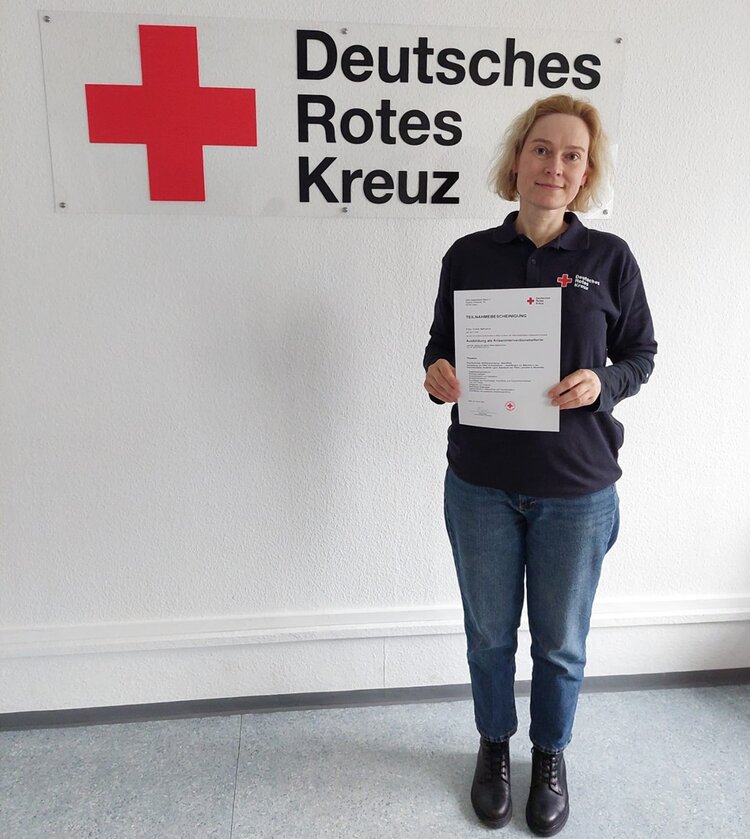 Frau Yuliia Nahorna steht in blauen Jeans, dunklen Chelsea Boots und dunklem Pullover lächelnd vor einem großen Schild des Deutschen Roten Kreuzes und hält ihre Teilnahmebescheinigung in die Kamera.