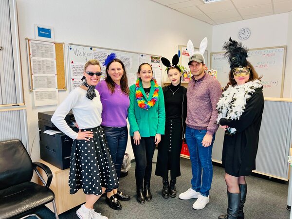 Faschingsdienstag an der Euro Akademie und den Euro-Schulen Dresden, Meißen und Riesa Eine Gruppe von sechs Personen in bunten, kreativen Kostümen posiert in einem Büro vor einem Whiteboard mit verschiedenen Notizen. Einige tragen Hasenohren, eine Person hat eine Federboa und eine Maske, während eine andere im Rockabilly-Stil mit Sonnenbrille und gepunktetem Rock gekleidet ist.