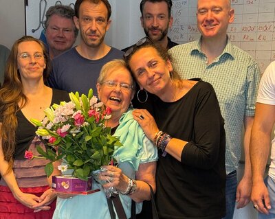 Eine fröhliche Gruppe von Menschen posiert in einem Raum vor einem Whiteboard. Im Vordergrund steht eine ältere Frau mit Blumenstrauß und einer Schachtel Pralinen, sie wird herzlich von einer anderen Frau umarmt. Die Gruppe lächelt in die Kamera.