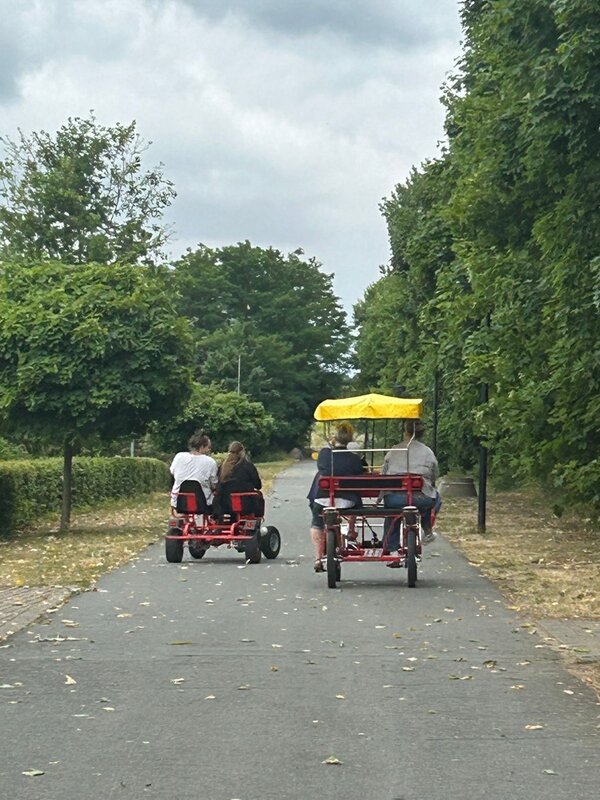 Die Auszubildenden sitzen auf zwei geliehenen Tretmobilen auf vier Rädern, mit mehreren Sitzplätzen. Eines der Tretmobile ist mit einem gelben Tuch überdacht.