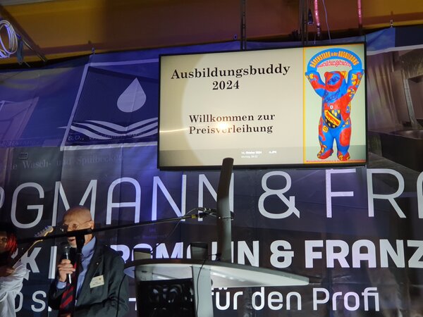 Reinickendorfer Ausbildungsbuddy geht an Netzwerkpartner der Euro-Schulen Berlin Ein Bühne mit grauem Rednerpult und ein Bildschirm im Hintergrund mit der Aufschrift Ausbildungsbuddy 2024.