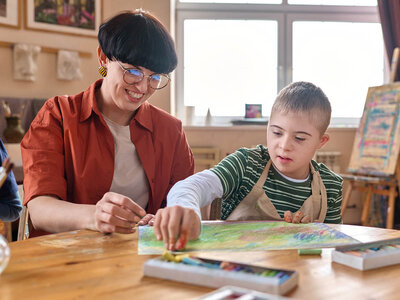 Inklusionsfachkraft Schwerpunkt "Schulassistenz und -begleitung" nach den Standards der Region Hannover Eine Frau mit kurzen Haaren und Brille sitzt lächelnd an einem Tisch und hilft einem Jungen mit Down-Syndrom beim Malen. Der Junge trägt ein gestreiftes Shirt und eine Schutzschürfe. Beide arbeiten gemeinsam an einem Bild mit bunten Pastellkreiden. Auf dem Tisch liegen weitere Kreiden und eine offene Kreidebox. Im Hintergrund sind Kunstutensilien und ein Fenster zu sehen.